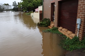 A flooded residential area with water covering the street and reaching up to the garages of brick-faced houses. Sandbags are placed in front of the doors to block water entry. Trees and shrubs are visible drenched in water, and the sky appears overcast, indicating ongoing or recent heavy rainfall.