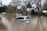 A silver car is partially submerged in floodwaters, surrounded by brown, murky water. Trees with bare branches and buildings with metal roofing are present in the background.