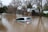 A silver car is partially submerged in floodwaters, surrounded by brown, murky water. Trees with bare branches and buildings with metal roofing are present in the background.