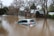 A silver car is partially submerged in floodwaters, surrounded by brown, murky water. Trees with bare branches and buildings with metal roofing are present in the background.