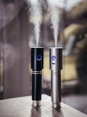 Two cylindrical electronic devices, one black and one silver, emit streams of vapor into the air. Both devices feature a glowing blue ring near the top and are placed on a wooden surface with a blurred background.