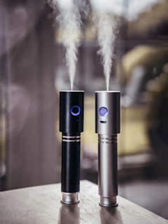 Two cylindrical electronic devices, one black and one silver, emit streams of vapor into the air. Both devices feature a glowing blue ring near the top and are placed on a wooden surface with a blurred background.