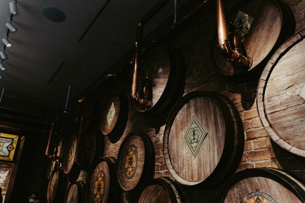 A row of large wooden barrels is mounted on a brick wall in a dimly lit room. The barrels have intricate designs on them, and copper pipes run over and around them, suggesting a brewing or distillery setting. Ambient lighting from above casts shadows, adding to the rustic and industrial aesthetic.