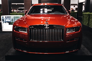 A luxurious red car showcased indoors with a prominent grille and sleek design elements. The setting includes dim lighting and reflections on the car's surface, with additional cars in the background and some greenery decor.