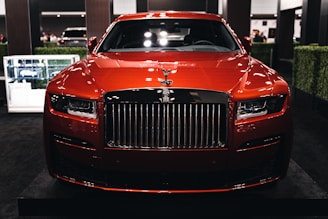 A luxurious red car showcased indoors with a prominent grille and sleek design elements. The setting includes dim lighting and reflections on the car's surface, with additional cars in the background and some greenery decor.
