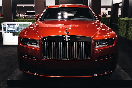 A luxurious red car showcased indoors with a prominent grille and sleek design elements. The setting includes dim lighting and reflections on the car's surface, with additional cars in the background and some greenery decor.
