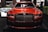 A luxurious red car showcased indoors with a prominent grille and sleek design elements. The setting includes dim lighting and reflections on the car's surface, with additional cars in the background and some greenery decor.