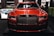 A luxurious red car showcased indoors with a prominent grille and sleek design elements. The setting includes dim lighting and reflections on the car's surface, with additional cars in the background and some greenery decor.