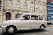 A white taxi is moving along a city street, with a blurred background consisting of ornate buildings. The architecture features detailed facades, including arches and windows. One of the buildings is covered with scaffolding on the right.