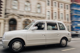 A white taxi is moving along a city street, with a blurred background consisting of ornate buildings. The architecture features detailed facades, including arches and windows. One of the buildings is covered with scaffolding on the right.