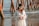 A pregnant woman stands by the sea, wearing a long, flowing white dress and a floral headband. Her hands are gently resting on her belly, forming a heart shape. In the background, wooden posts from a pier extend into the water, with gentle waves in view.