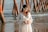 A pregnant woman stands by the sea, wearing a long, flowing white dress and a floral headband. Her hands are gently resting on her belly, forming a heart shape. In the background, wooden posts from a pier extend into the water, with gentle waves in view.