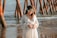 A pregnant woman stands by the sea, wearing a long, flowing white dress and a floral headband. Her hands are gently resting on her belly, forming a heart shape. In the background, wooden posts from a pier extend into the water, with gentle waves in view.