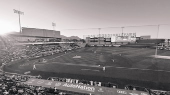 A baseball game is taking place in a stadium filled with spectators. The players are positioned on the field, which is surrounded by advertising banners. The setting sun in the background casts a warm glow over the scene.