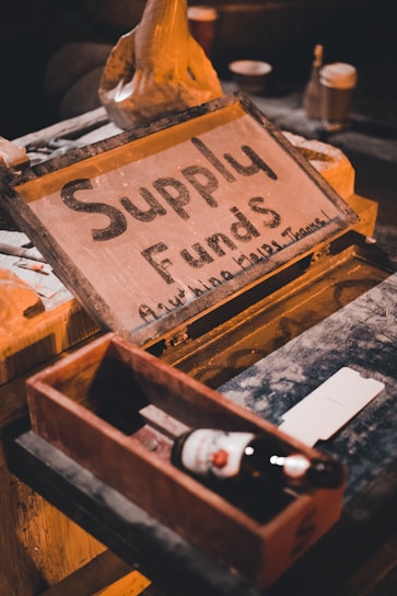 A rustic setup on a wooden table features a chalkboard sign with the words 'Supply Funds' written on it. Nearby, a wooden box holds a single beer bottle, and a few other objects such as jars and containers are visible in the dim, warm lighting.