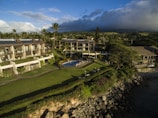 Aerial view of a coastal resort featuring multiple buildings with solar panels on rooftops, surrounded by lush greenery and a well-maintained lawn. A swimming pool is centrally located with people lounging around it. The area is bordered by a rocky shoreline with the ocean partially visible on the right.