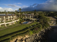 Aerial view of a coastal resort featuring multiple buildings with solar panels on rooftops, surrounded by lush greenery and a well-maintained lawn. A swimming pool is centrally located with people lounging around it. The area is bordered by a rocky shoreline with the ocean partially visible on the right.