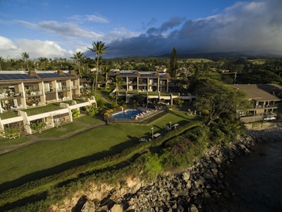 Aerial view of a coastal resort featuring multiple buildings with solar panels on rooftops, surrounded by lush greenery and a well-maintained lawn. A swimming pool is centrally located with people lounging around it. The area is bordered by a rocky shoreline with the ocean partially visible on the right.