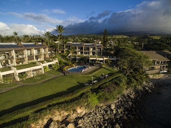 Aerial view of a coastal resort featuring multiple buildings with solar panels on rooftops, surrounded by lush greenery and a well-maintained lawn. A swimming pool is centrally located with people lounging around it. The area is bordered by a rocky shoreline with the ocean partially visible on the right.