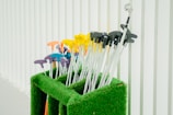 A collection of mini-golf putters with colorful handles is neatly arranged in a green artificial grass holder. The handles are in various colors, including yellow, orange, purple, and blue, and are set against a background of vertical white panels.