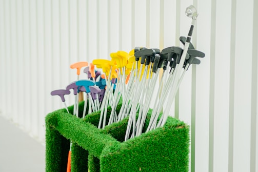 A collection of mini-golf putters with colorful handles is neatly arranged in a green artificial grass holder. The handles are in various colors, including yellow, orange, purple, and blue, and are set against a background of vertical white panels.