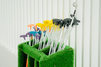 A collection of mini-golf putters with colorful handles is neatly arranged in a green artificial grass holder. The handles are in various colors, including yellow, orange, purple, and blue, and are set against a background of vertical white panels.
