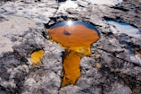 A rocky surface with irregular puddles of water, one of which prominently reflects sunlight with a bright starburst effect. The water appears to have various earthy tones, ranging from light orange to darker hues, surrounded by weathered and eroded rock formations.