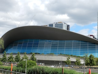 A modern architectural building featuring an expansive curved roof and a facade composed of large blue-tinted glass panels. The foreground includes lush green trees and plants, while the background has tall cranes and additional buildings under a partly cloudy sky.