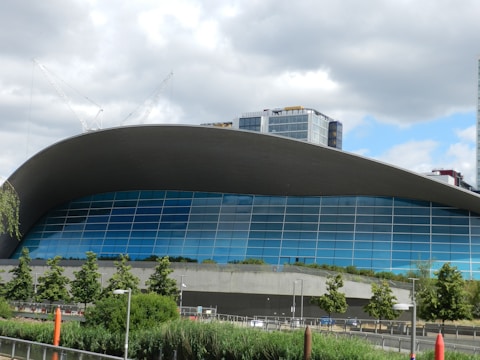 A modern architectural building featuring an expansive curved roof and a facade composed of large blue-tinted glass panels. The foreground includes lush green trees and plants, while the background has tall cranes and additional buildings under a partly cloudy sky.