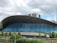A modern architectural building featuring an expansive curved roof and a facade composed of large blue-tinted glass panels. The foreground includes lush green trees and plants, while the background has tall cranes and additional buildings under a partly cloudy sky.