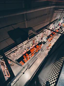 An overhead view of a retail store interior with industrial design features, showcasing multiple shelves filled with merchandise. The store has a modern aesthetic with a mix of metal structures and bright lighting. Numerous orange boxes line the shelves, adding a splash of color to the neutral space. Several shoppers, including one wearing a yellow shirt, are visible browsing the aisles.