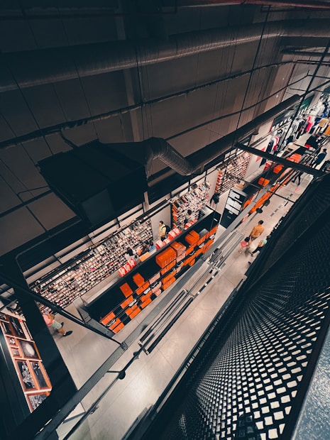 An overhead view of a retail store interior with industrial design features, showcasing multiple shelves filled with merchandise. The store has a modern aesthetic with a mix of metal structures and bright lighting. Numerous orange boxes line the shelves, adding a splash of color to the neutral space. Several shoppers, including one wearing a yellow shirt, are visible browsing the aisles.