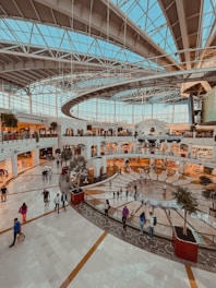 A spacious indoor shopping mall with a large glass ceiling allowing natural light to flood the area. The multilevel structure has numerous shops with bright storefronts, scattered potted trees, and groups of people walking and socializing. The architecture features modern design elements with a mix of straight and curved lines.