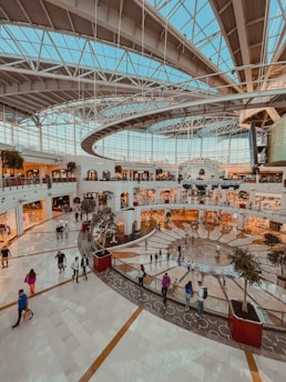 A spacious indoor shopping mall with a large glass ceiling allowing natural light to flood the area. The multilevel structure has numerous shops with bright storefronts, scattered potted trees, and groups of people walking and socializing. The architecture features modern design elements with a mix of straight and curved lines.