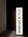 A vertically oriented lightbox displays the text 'DREAMS HAVE NO TITLES' in bold, black uppercase letters on a white background. The lightbox is positioned in a dimly lit space, creating a stark contrast between the illuminated sign and the dark surroundings.