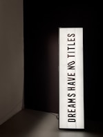 A vertically oriented lightbox displays the text 'DREAMS HAVE NO TITLES' in bold, black uppercase letters on a white background. The lightbox is positioned in a dimly lit space, creating a stark contrast between the illuminated sign and the dark surroundings.