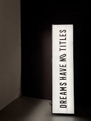 A vertically oriented lightbox displays the text 'DREAMS HAVE NO TITLES' in bold, black uppercase letters on a white background. The lightbox is positioned in a dimly lit space, creating a stark contrast between the illuminated sign and the dark surroundings.