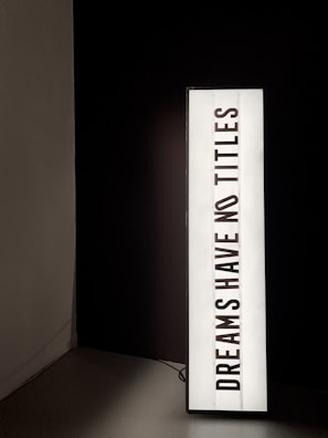 A vertically oriented lightbox displays the text 'DREAMS HAVE NO TITLES' in bold, black uppercase letters on a white background. The lightbox is positioned in a dimly lit space, creating a stark contrast between the illuminated sign and the dark surroundings.