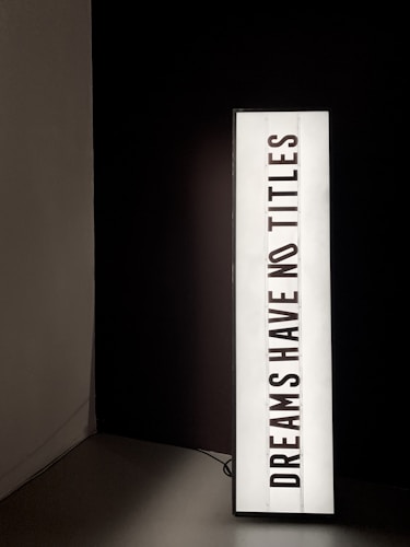 A vertically oriented lightbox displays the text 'DREAMS HAVE NO TITLES' in bold, black uppercase letters on a white background. The lightbox is positioned in a dimly lit space, creating a stark contrast between the illuminated sign and the dark surroundings.