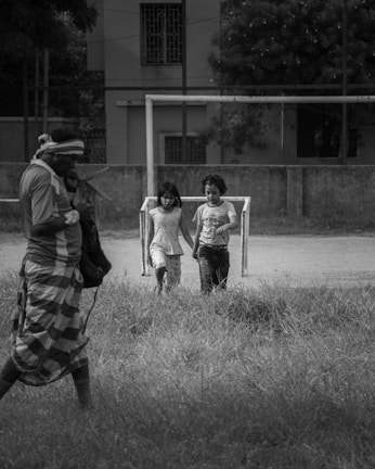 Two children walk hand in hand near a makeshift goalpost on a grassy field, while an adult stands nearby, carrying a bag and wearing a checked cloth wrap around the waist. The background features a building with barred windows and lush trees.