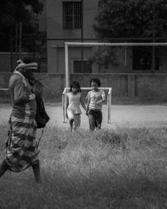 Two children walk hand in hand near a makeshift goalpost on a grassy field, while an adult stands nearby, carrying a bag and wearing a checked cloth wrap around the waist. The background features a building with barred windows and lush trees.