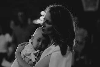 A woman holds a baby close in a tender embrace, creating a sense of intimacy and care. The image is in black and white, emphasizing shadows and highlights.