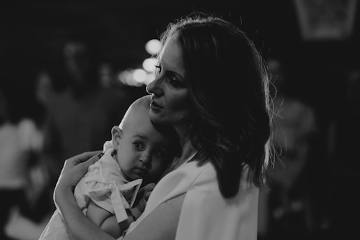A woman holds a baby close in a tender embrace, creating a sense of intimacy and care. The image is in black and white, emphasizing shadows and highlights.