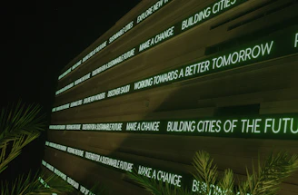 A modern digital display board features phrases promoting sustainability, future cities, and innovation. It emits a green glow against a dark background, while palm fronds partially frame the view.