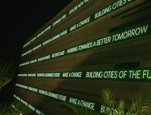 A modern digital display board features phrases promoting sustainability, future cities, and innovation. It emits a green glow against a dark background, while palm fronds partially frame the view.