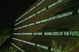 A modern digital display board features phrases promoting sustainability, future cities, and innovation. It emits a green glow against a dark background, while palm fronds partially frame the view.
