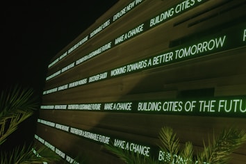 A modern digital display board features phrases promoting sustainability, future cities, and innovation. It emits a green glow against a dark background, while palm fronds partially frame the view.