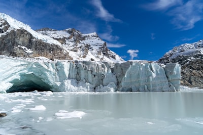 A large glacier is depicted, with icy formations extending into a frozen body of water. Snow-covered rocky mountains rise in the background under a vivid blue sky with some scattered clouds. The glacier has a large, dark cavernous entrance on the left side.
