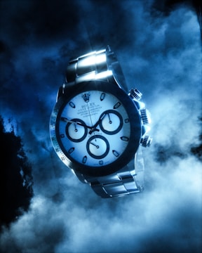 A luxurious watch with a metallic strap and detailed chronograph dials is prominently featured against a smoky and dramatic blue background. The sleek and polished surface of the watch reflects light, enhancing its sophisticated appearance.