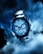 A luxurious watch with a metallic strap and detailed chronograph dials is prominently featured against a smoky and dramatic blue background. The sleek and polished surface of the watch reflects light, enhancing its sophisticated appearance.